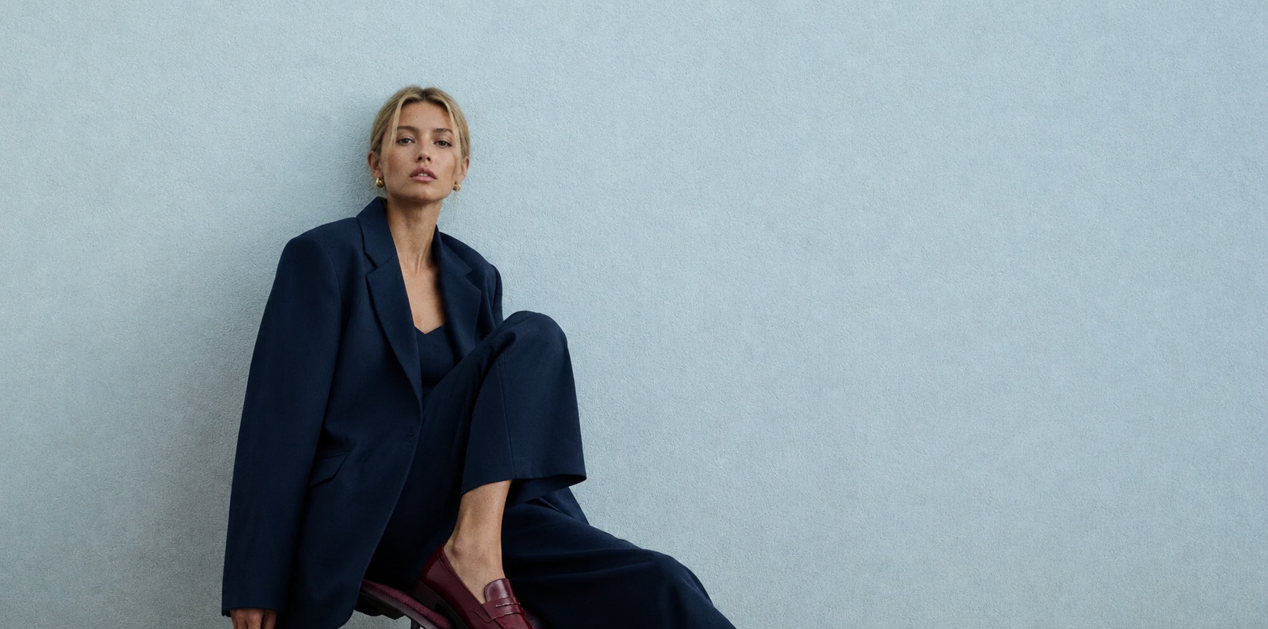Femme en tailleur sombre assise devant un mur bleu clair, campagne Lena Paris